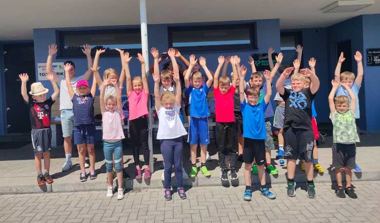 Gruppe von Kindern in Sportkleidung stehen mit erhobenen Händen vor einem Gebäude, sonniger Tag, fröhliche Stimmung.