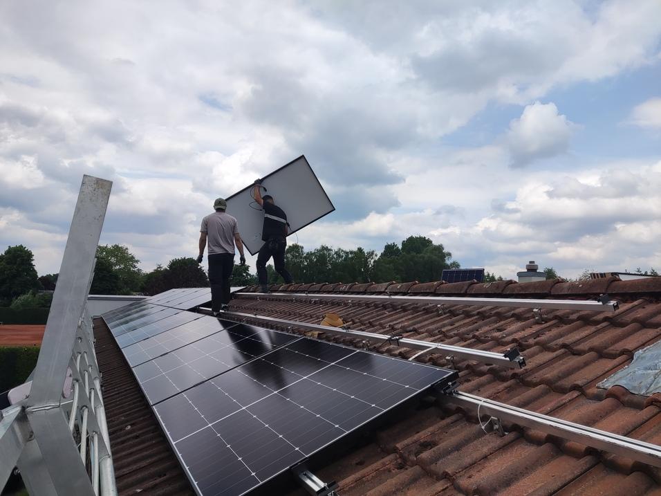 Zwei Arbeiter installieren Solarpanels auf einem Dach mit roten Ziegeln unter bewölktem Himmel.