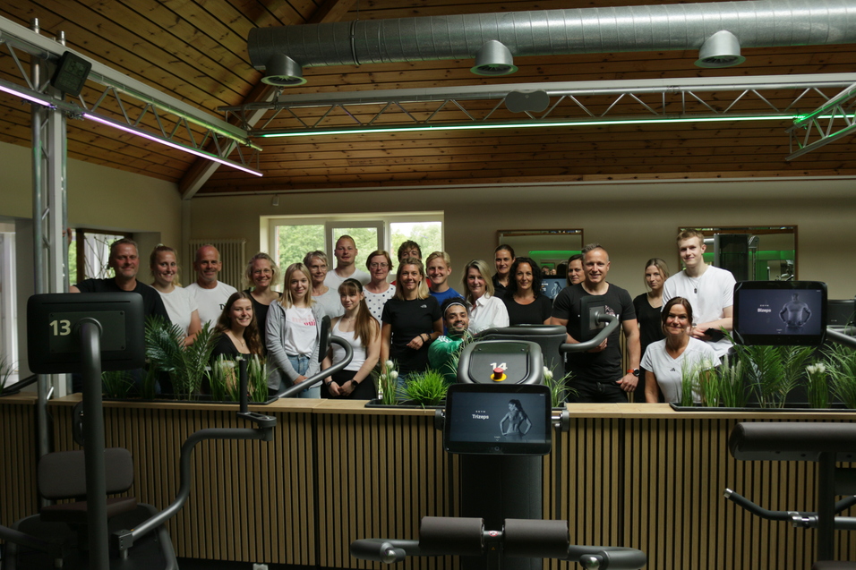 Gruppe von 20 Personen in einem Fitnessstudio, umgeben von Trainingsgeräten und Pflanzen, vor Holzvertäfelung.