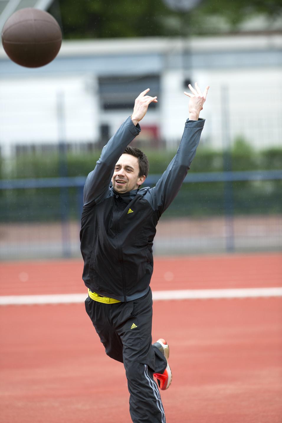 Mann in Sportbekleidung wirft einen Fußball auf einem roten Sportplatz unter bewölktem Himmel.