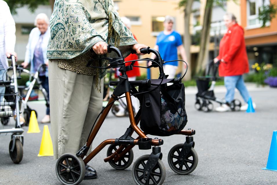 Nahaufnahme einer Person mit einem Rollator, umgeben von weiteren Senioren, die auf einem Außenbereich üben.
