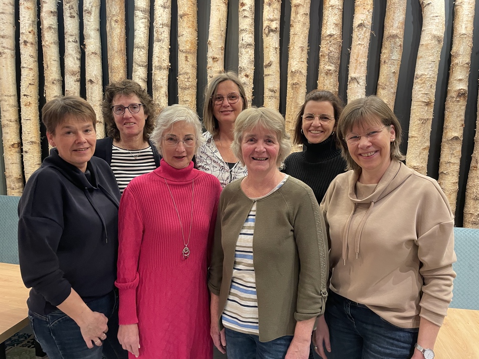 Gruppe von sieben Frauen lächelnd vor einer Wand aus Baumstämmen, in einem informellen Innenraum.