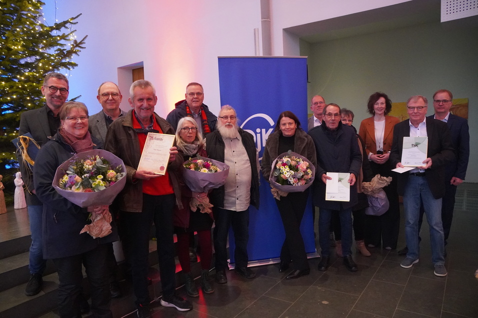 Gruppe von Menschen mit Blumensträußen und Urkunden vor einem blauen Banner in einem festlich geschmückten Raum.