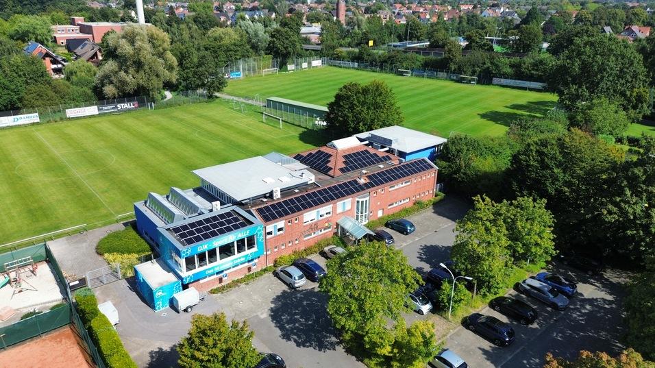 Luftaufnahme eines Sportgeländes mit Fußballfeld und einem Gebäude mit Solarpanelen und Parkplatz.