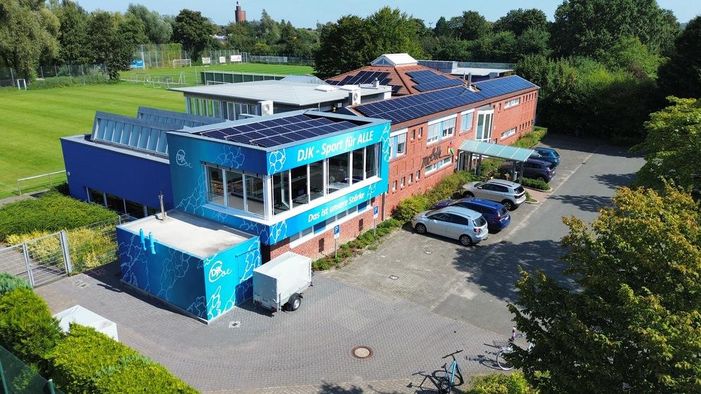 Blick auf ein Sportzentrum mit Solarzellen, umgeben von Gr&uuml;nfl&auml;chen und Autos auf einem Parkplatz.