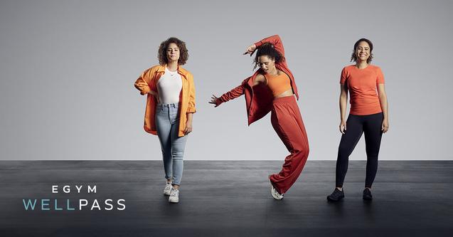 Drei Frauen in Sportkleidung posieren auf einem grauen Hintergrund, eine tanzt, die anderen stehen entspannt.