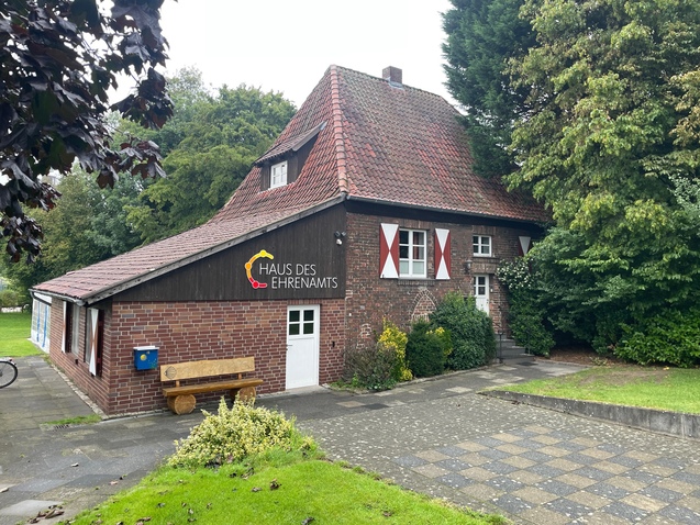 Haus des Ehrenamts, ein zweigeschossiges Gebäude mit rotem Satteldach und Holzfassade, umgeben von Bäumen und Grünflächen.