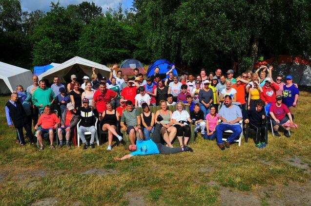 Gruppenfoto von 65 Personen im Freien, umgeben von Zelten und Bäumen, einige winken und lachen.