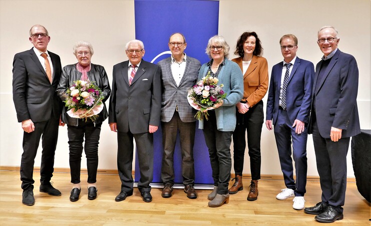 Gruppenfoto von sieben Personen mit Blumensträußen, stehend vor einem blauen Banner in einem Veranstaltungsraum.