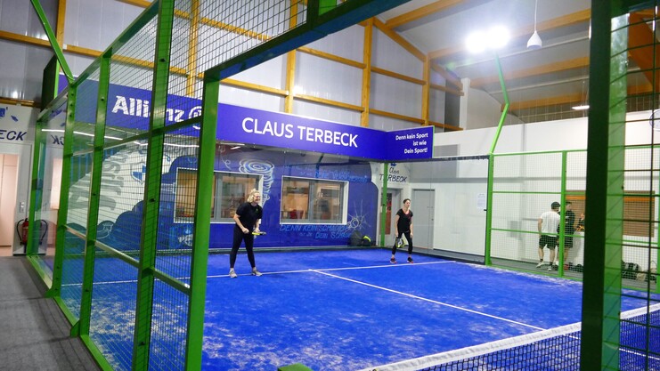 Zwei Spielerinnen stehen auf einem blauen Padelplatz, umgeben von einem grünen Netz und einer beschrifteten Wand.