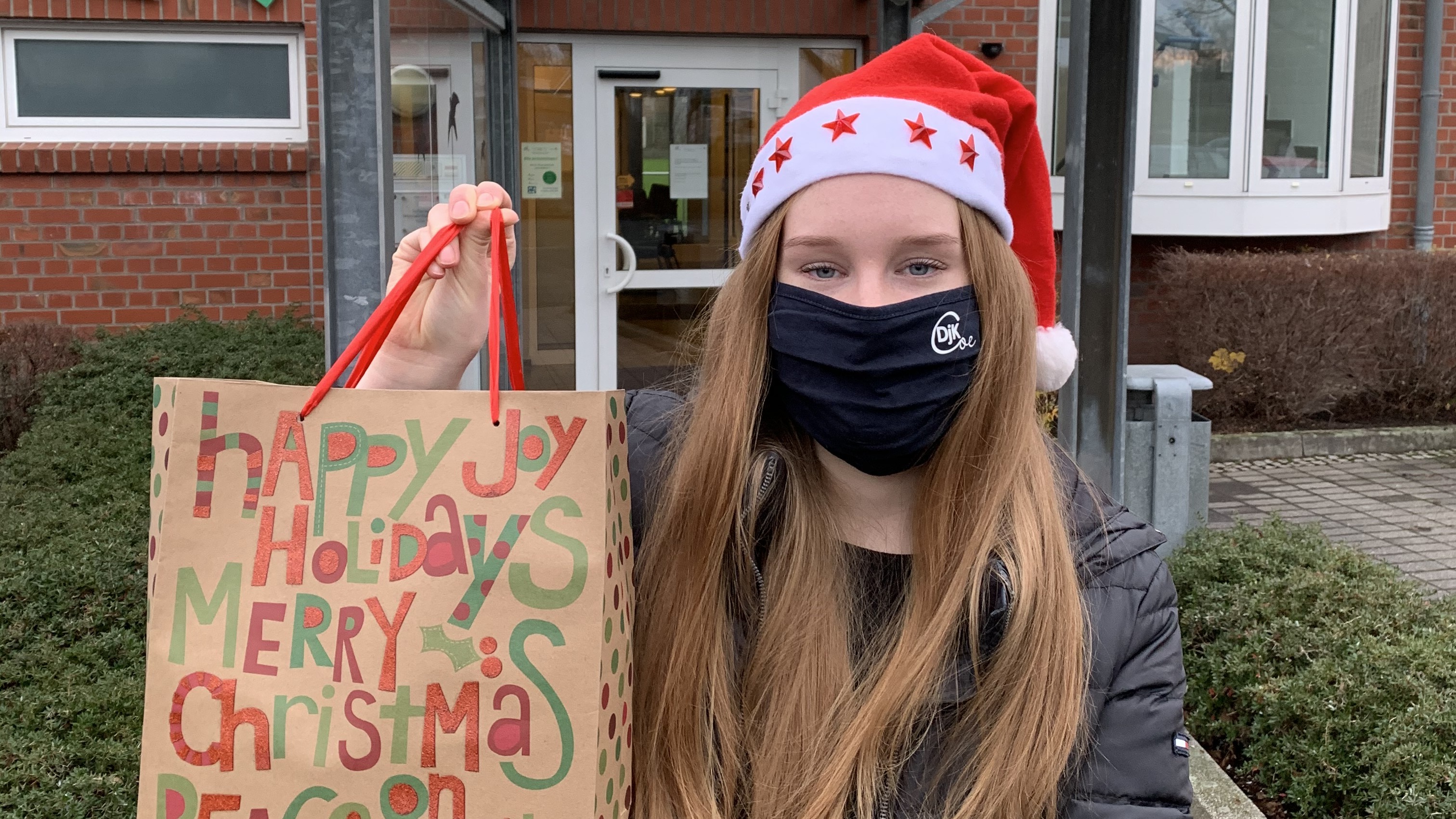 Junge mit roter Weihnachtsmütze hält eine bunt beschriftete Geschenktüte vor einem Gebäude im Freien.