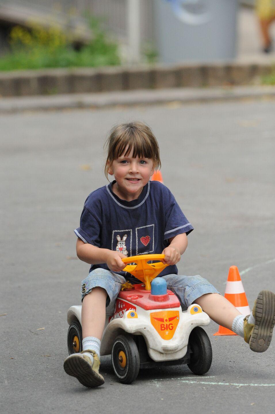 Lächelndes Kind fährt auf einem kleinen roten Spielauto zwischen Kegeln auf einem Asphaltweg.