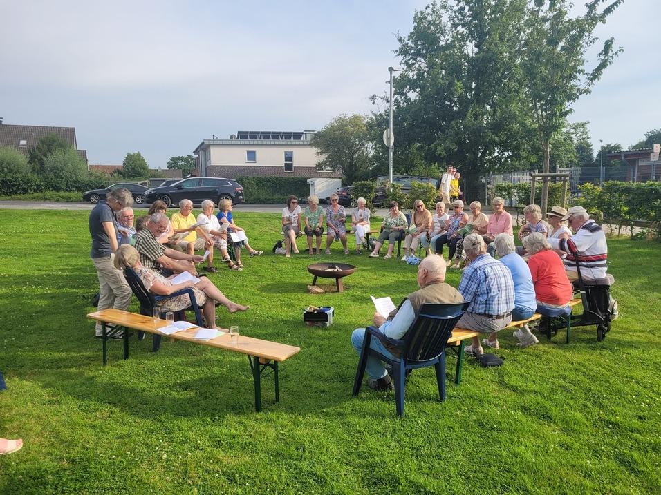 Gruppe von Senioren sitzt im Freien auf Stühlen und Bänken um ein Feuer, umgeben von Grünflächen und Bäumen.
