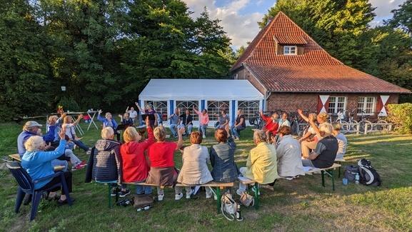 Gruppe von Menschen sitzt vor einem Haus, während eine Person im Vordergrund eine Aktivität leitet. Bühne im Freien.