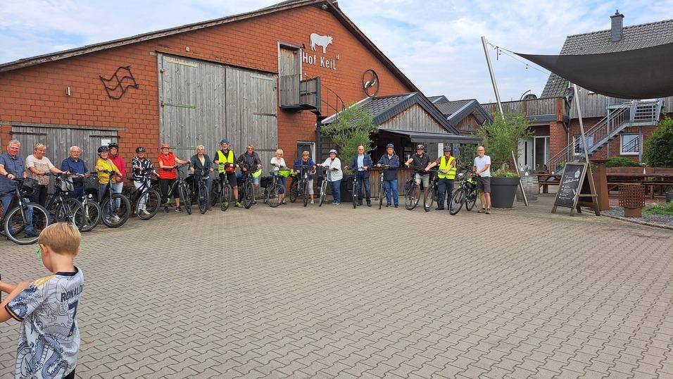 Gruppe von Radfahrern mit Fahrrädern vor einem Bauernhof, einige tragen Helme und reflektierende Westen.