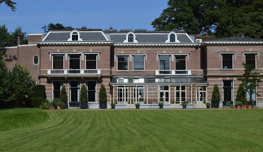 Historisches Backsteinhaus mit großen Fenstern, umgeben von Rasen und Bäumen im Freien.