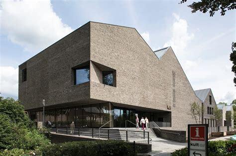 Moderne, dreieckige Architektur eines Gebäudes aus Klinker mit großen Fenstern und umliegendem Grün.