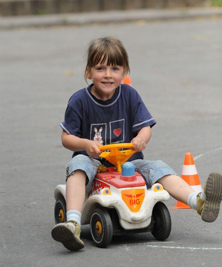 Lächelndes Kind fährt auf einem kleinen roten Spielauto zwischen Kegeln auf einem Asphaltweg.