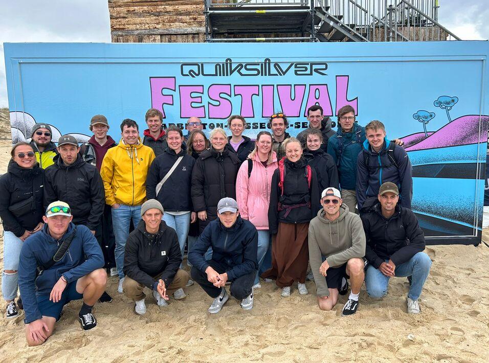 Gruppe von 20 Personen vor einer bunten Festivalwand, mit Sandboden im Hintergrund und Baugerüst hinter ihnen.
