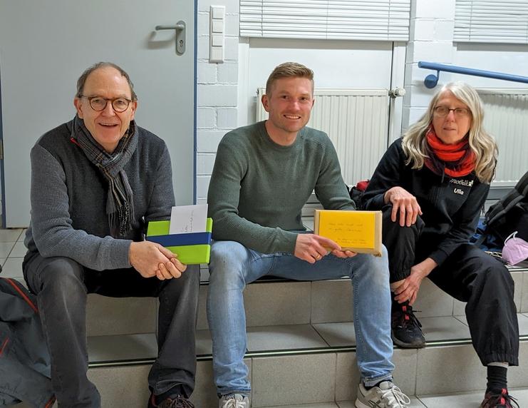 Drei Personen sitzen auf einer Treppe, halten Geschenke und lachen in einem hellen Raum mit weißen Wänden.
