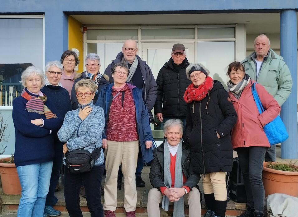 Gruppe von zwölf Menschen steht vor einem Gebäude, einige lächeln, während andere neutral schauen.