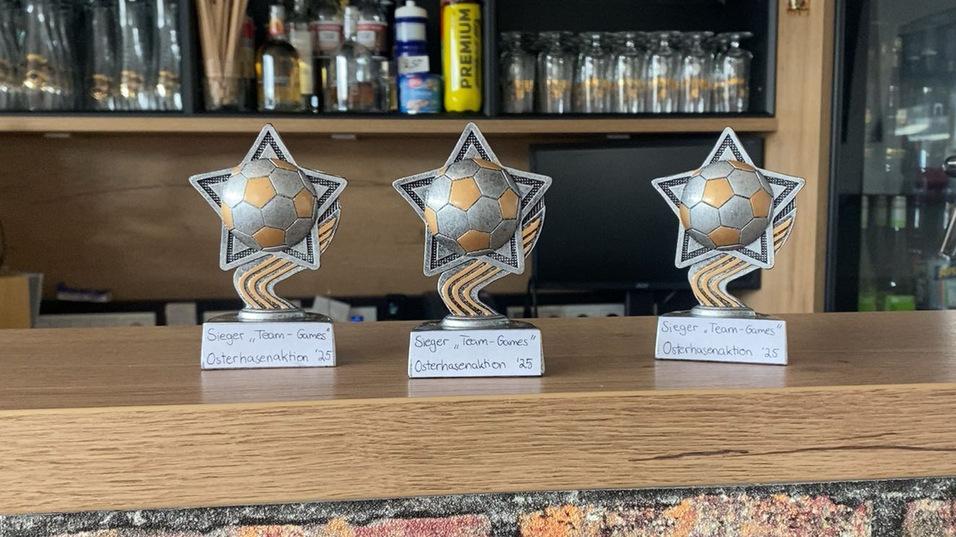 Drei Fußballtrophäen mit goldenen und silbernen Elementen stehen auf einem Holztisch in einer Bar.