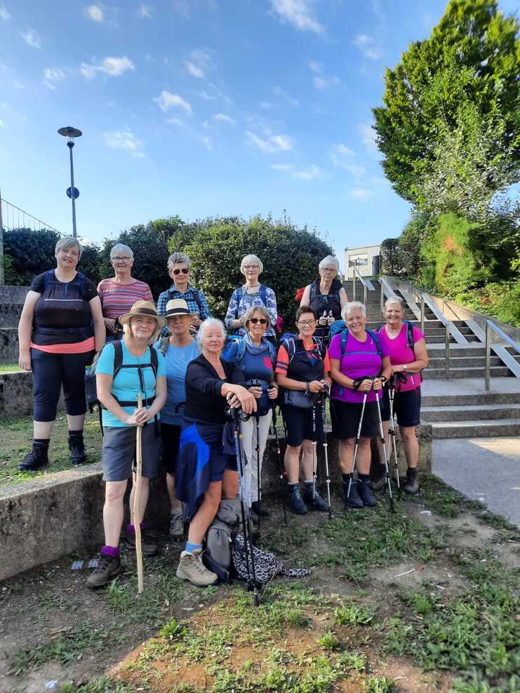 Gruppe von 12 Frauen mit Wanderstöcken steht lächelnd auf einer Treppe vor grüner Vegetation und blauem Himmel.