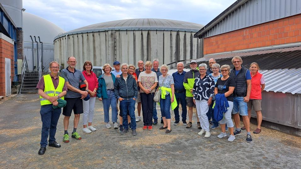 Gruppe von 20 Personen in Arbeitskleidung, stehend im Freien vor einer Biogasanlage mit großen, runden Behältern.