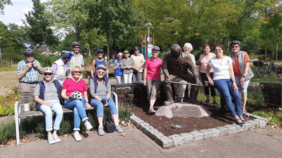 Gruppe von Radfahrern in Helmen posiert neben einer Statue in einem Park. Hintergrund mit Bäumen und Wiese.