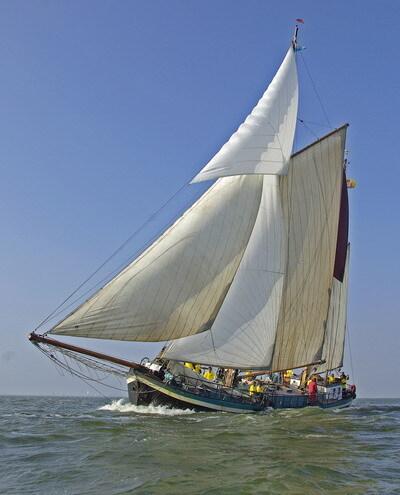 Ein Segelschiff mit großen weißen Segeln fährt bei sonnigem Wetter über das Wasser, Passagiere an Deck.