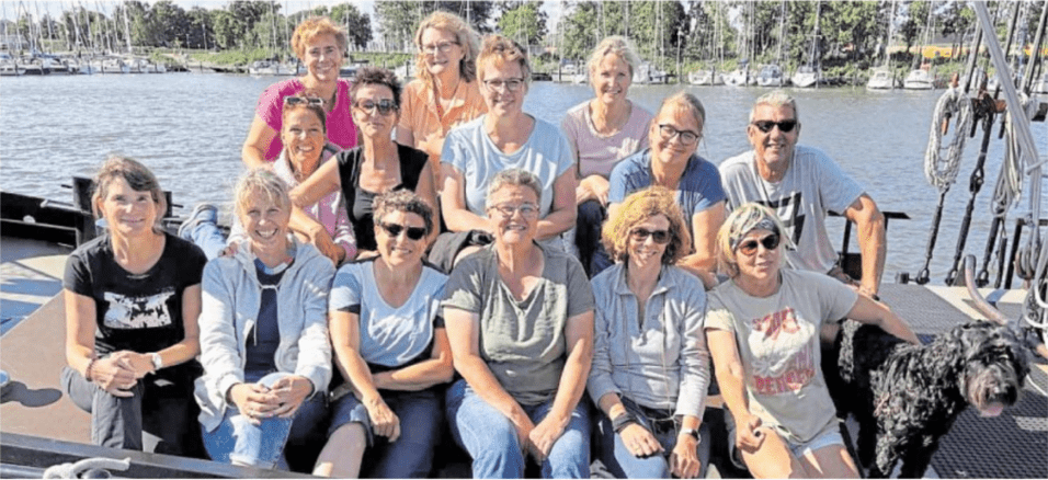 Gruppe von zwölf Personen und einem Hund sitzt an einer Hafenkante mit Segelbooten im Hintergrund.