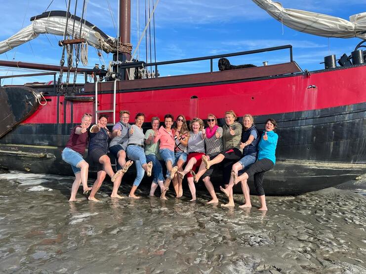 Gruppe von Frauen, die barfuß am Meer vor einem roten Schiff posieren und Daumen hoch zeigen.