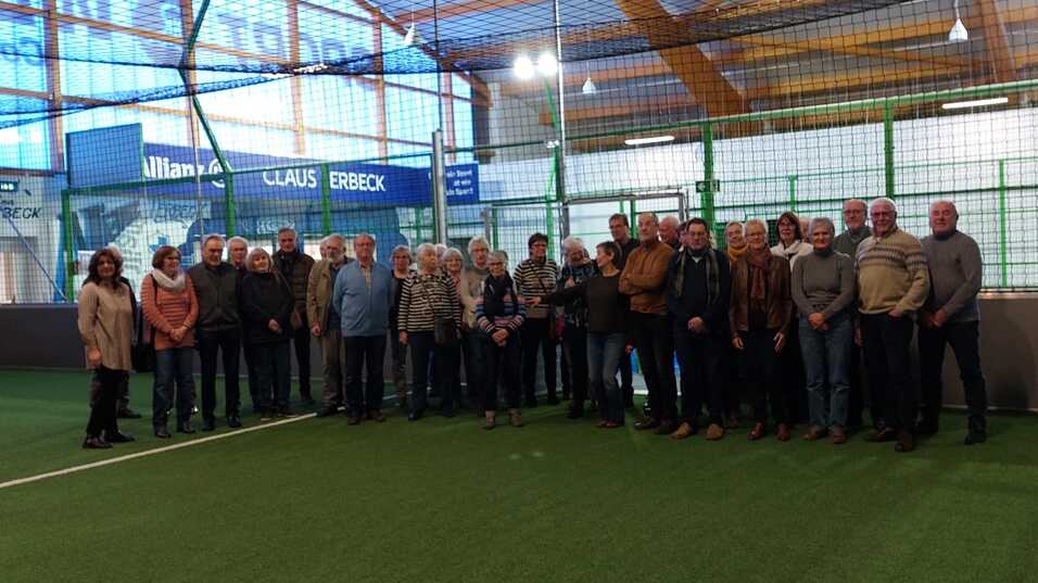 Gruppe von Menschen steht auf einem Kunstrasenfeld in einer Sporthalle, umgeben von großen Fenstern und einem Netz.