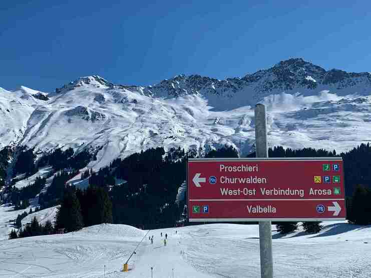 Skisprung-Schild mit Wegweisern zu Proscheri, Churwalden, Arosa und Valbella vor schneebedeckten Bergen.