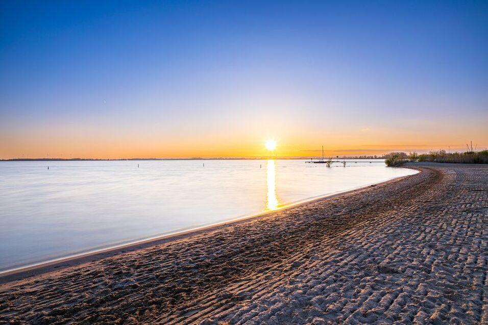 Sonnenuntergang über einem ruhigen See, mit feinem Sandstrand und reflektierendem Wasser im Vordergrund.