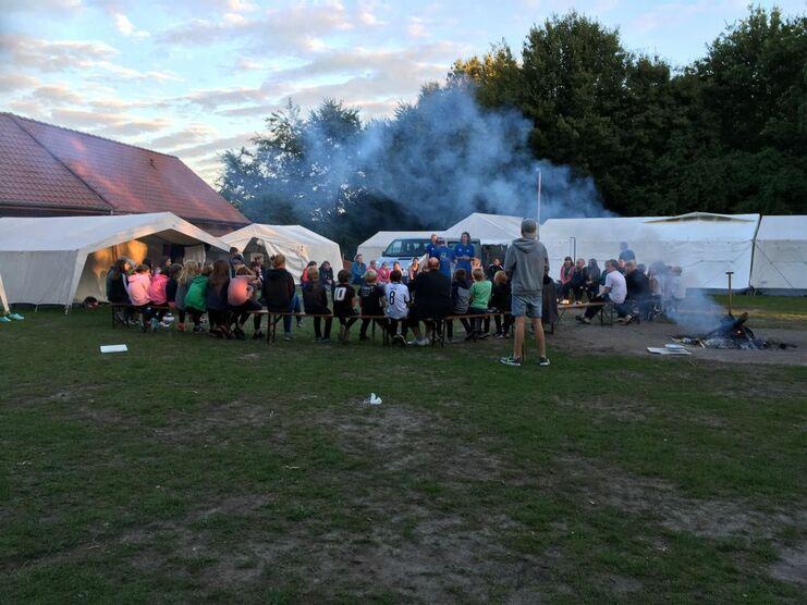 Gruppensitzung am Lagerfeuer mit Kindern und Betreuern vor Zelten auf einer Wiese. Rauch steigt in den Abendhimmel.