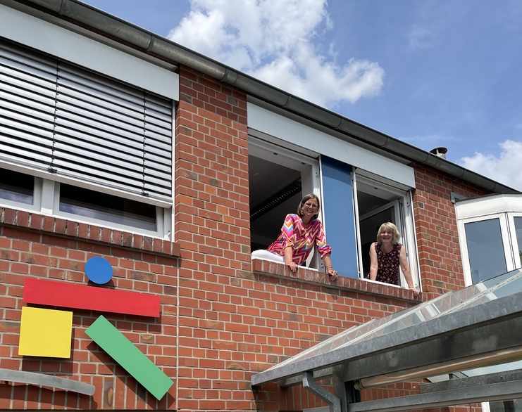 Zwei Frauen lehnen aus einem Fenster eines Ziegelgebäudes, darunter bunte geometrische Formen aus Holz.