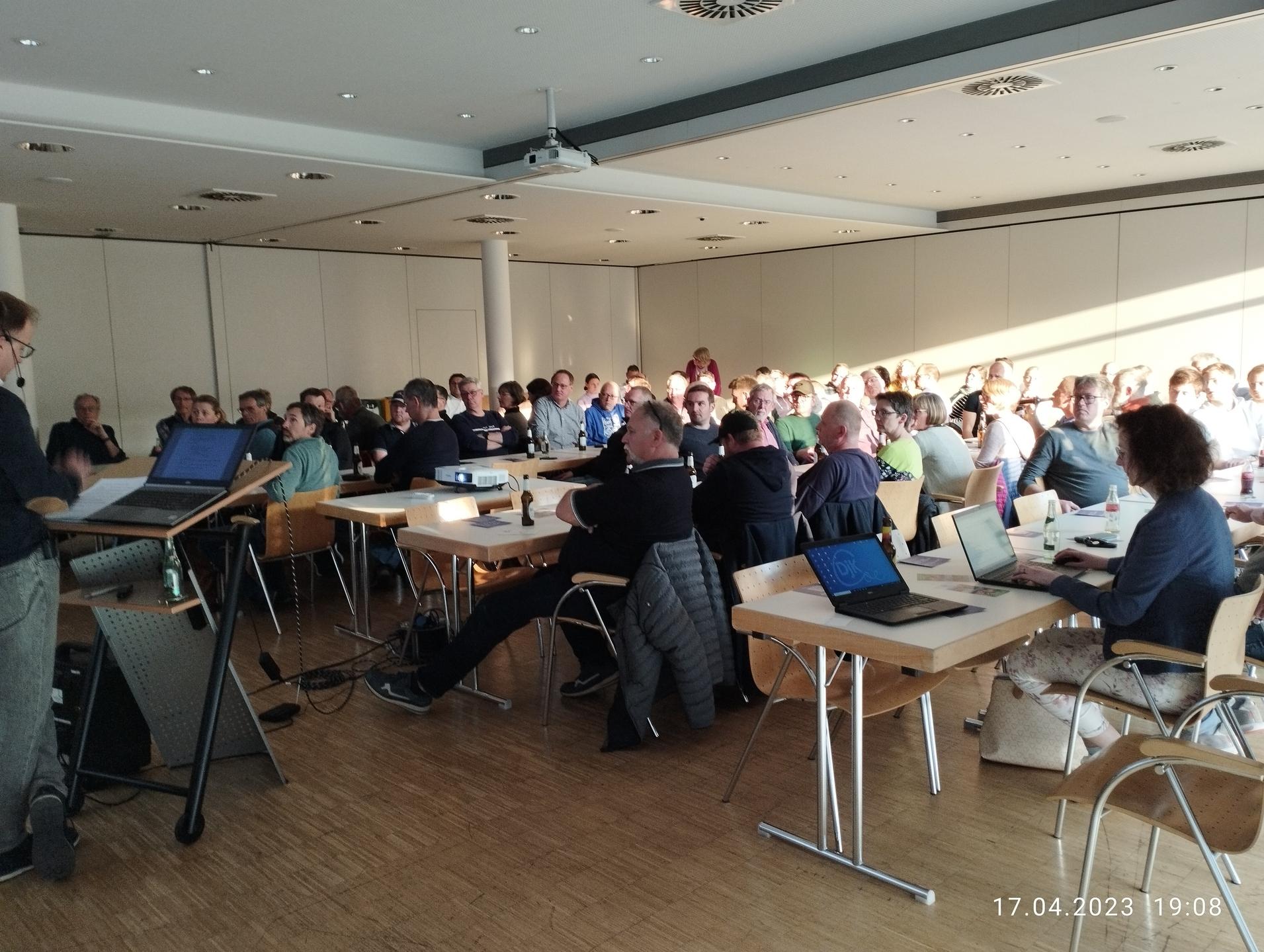 Teilnehmer sitzen in einem hellen Raum bei einer Präsentation, ein Referent steht vorne am Tisch mit Laptops.