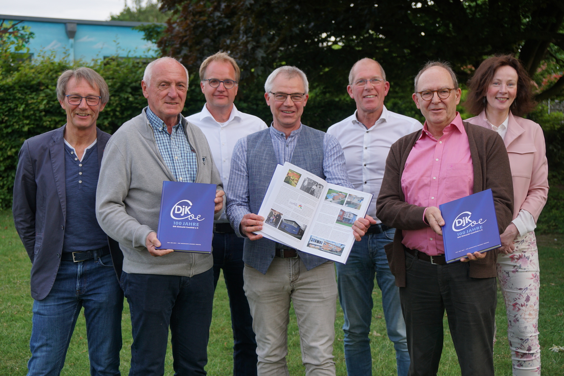 Gruppe von sieben Personen posiert im Freien und hält Jubiläumsbroschüren der DJK in der Hand.