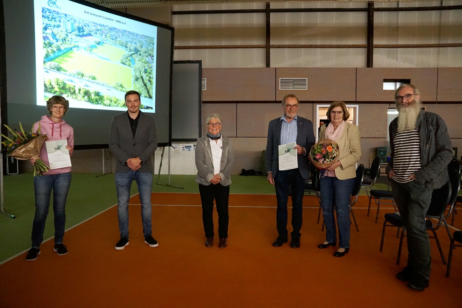Gruppenszene bei einer Ehrungsveranstaltung mit fünf Personen, die Urkunden und Blumen präsentieren, vor einer Präsentation.