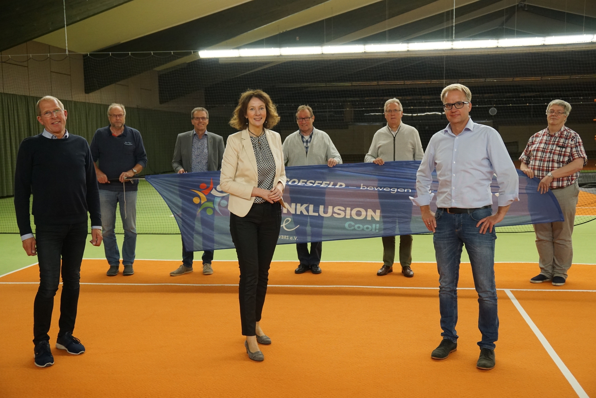 Gruppenteffen auf einem Tennisplatz mit Banner für „Inklusion“ und Unterstützern, die zum Event stehen.