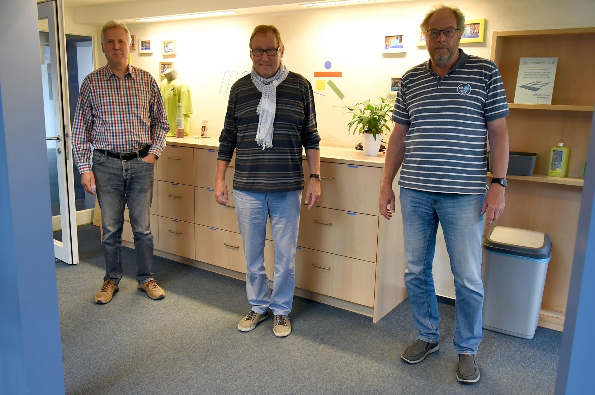 Drei Männer stehen in einem Büro vor einem Holztresen mit Pflanzen und Bildern im Hintergrund, die Wände sind blau.