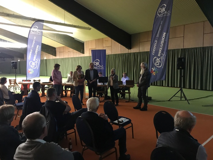 Veranstaltung in einer Sporthalle mit Personen, die an einem Rednerpult stehen, und Publikum in Sitzreihen.