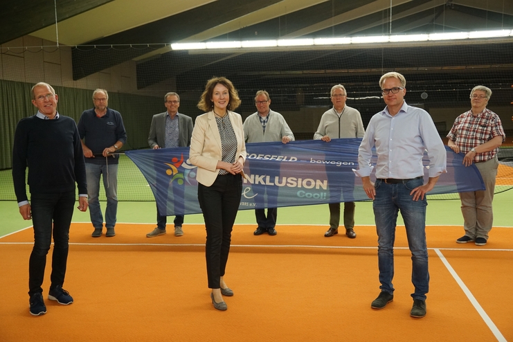 Gruppenteffen auf einem Tennisplatz mit Banner für „Inklusion“ und Unterstützern, die zum Event stehen.