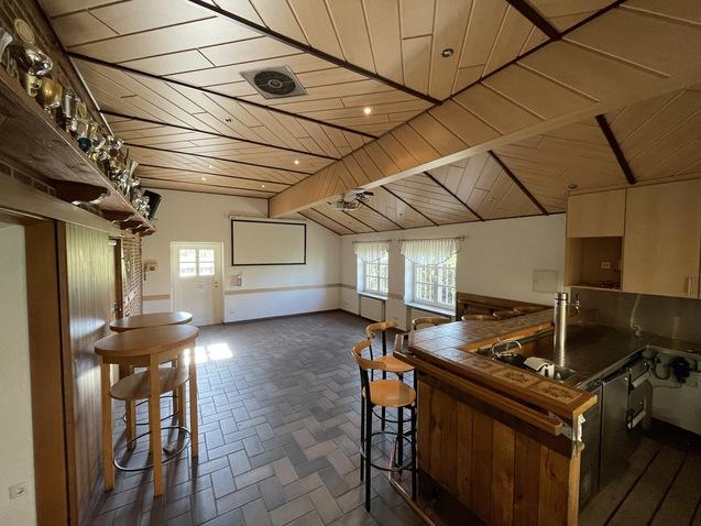 Geräumiger Veranstaltungsraum mit Holzdecke, Bar, Tisch und Fenster für Tageslicht. Pokale an der Wand sichtbar.