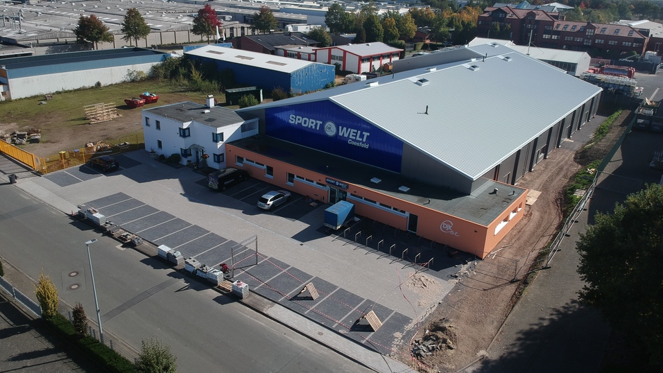 Blick auf ein modernes Sportgebäude mit Parkplätzen, umgeben von Grünflächen und Industrieanlagen im Hintergrund.