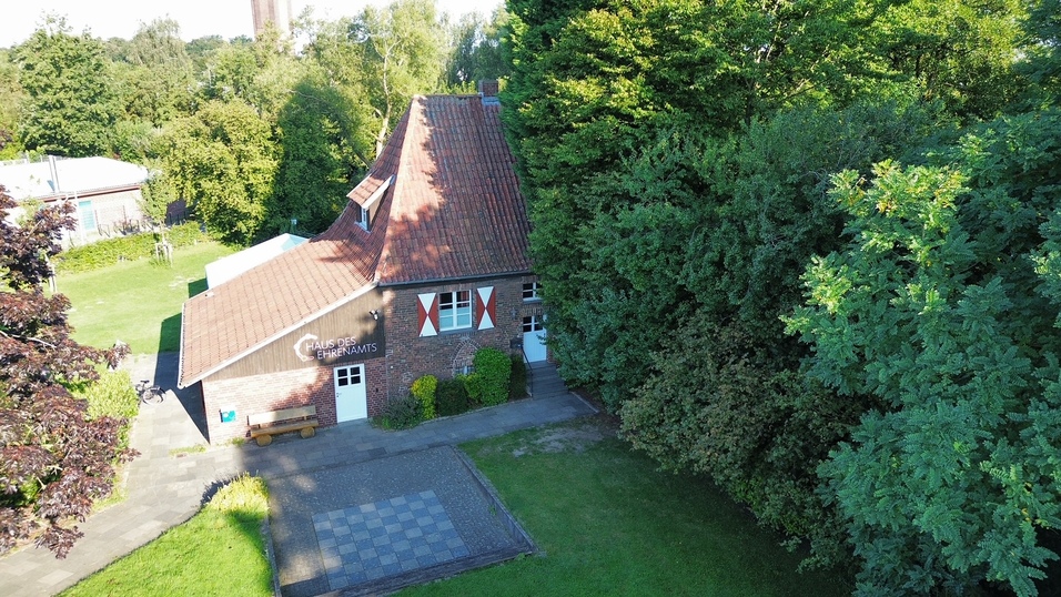 Luftaufnahme eines Hauses mit der Aufschrift „Haus des Ehrenamts“, umgeben von Bäumen und einer gepflasterten Fläche.
