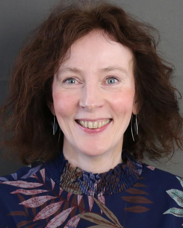 Frau mit lockigen, braunen Haaren und einem grauen Blazer l&auml;chelt vor einem bunten Hintergrund.