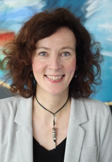 Frau mit lockigen, braunen Haaren und einem grauen Blazer lächelt vor einem bunten Hintergrund.