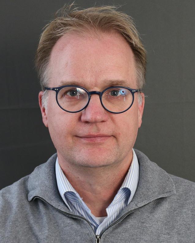 Portr&auml;t eines Mannes mit Brille und blauer Hemd vor einem Fenster mit wei&szlig;en Rahmen.